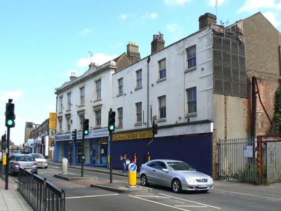 Two adjoining white painted brick-built properties, both of 3 storeys with ground floor shops, nearest property is boarded up