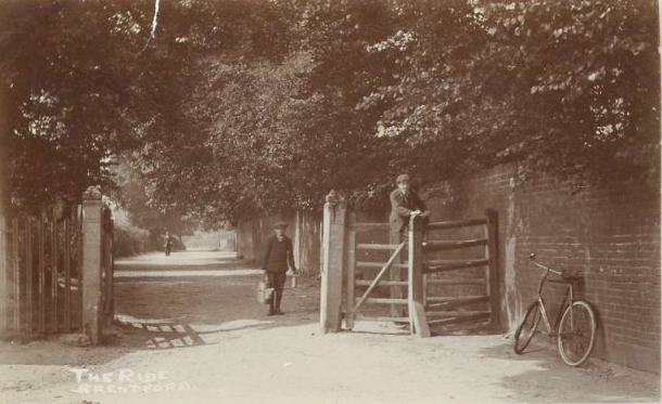 Sepia view of entry to lane, with stout wooden gate posts and a pedestrian entrance; the lane is bounded by a wall to the right