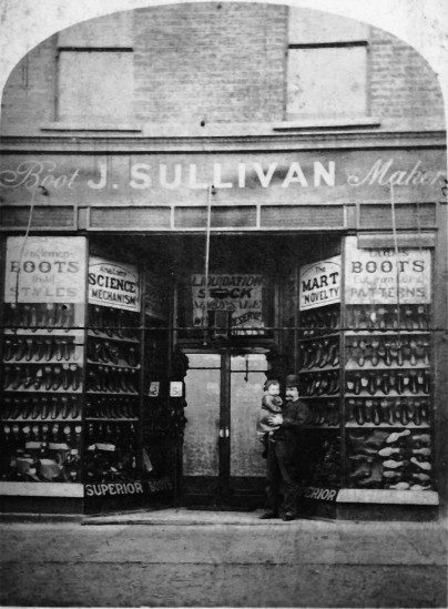 shop with recessed door and windows on either side, filled with rows of boots and shoes; in the doorway a smartly dressed man holds a small girl in his arms; the sign over the shop reads J. Sullivan, boot makers