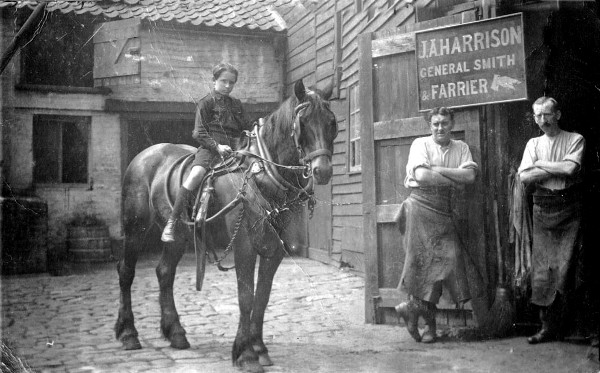 B/w photo showing corner of paved yard; boy on horse; two blacksmiths stand in doorway to smithy looking into the yard