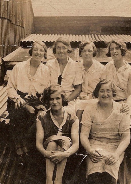 Photo taken outside, six cheerful ladies dressed in  summer clothes,  with backdrop of corrugated iron roofing