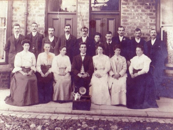 Group of 17 adults, more males, taken outside terrace house; gifts: clock and silver coffee pot on display