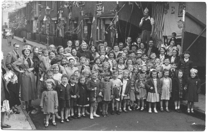Children and their mothers (and a few fathers) in a road of terraced houses bedecked with flags, streamers and bunting