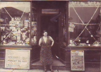 Double-fronted shop with recessed door with a lady; the windows have tinsel or streamers
