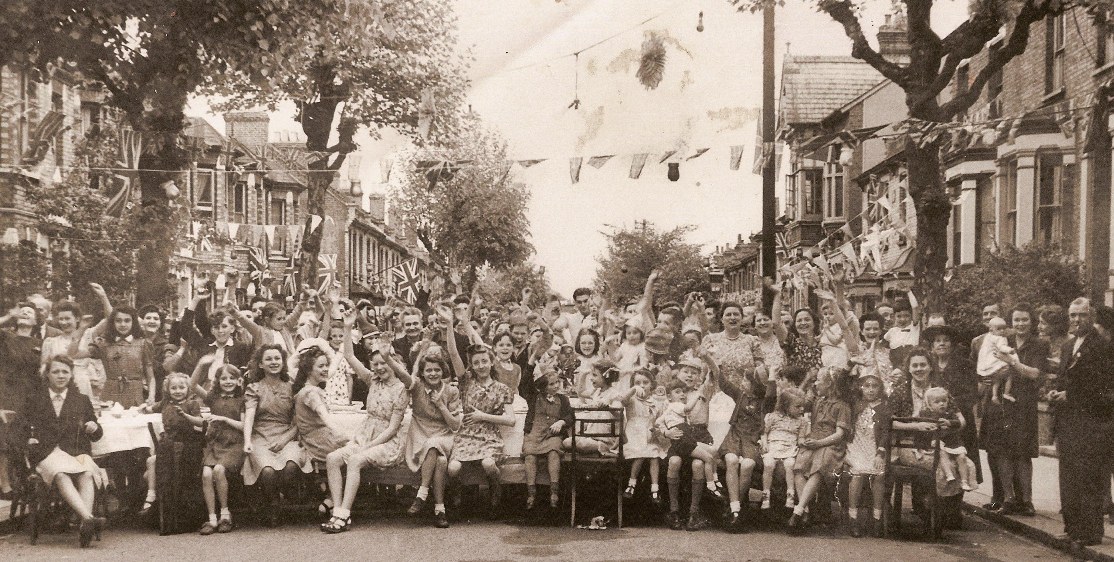Around 70 happy people, including lots of children and mothers, many in short sleeve dresses, waving at the camera.  They are sitting at a tableclothed table which runs across Hamilton Road, with many Union Jack flags and bunting. 