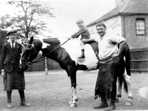 B/w photo, small boy on piebald horse whose head is held by an elderly man in a kilt; a cheery 40ish man with blacksmith's apron stands by the boy