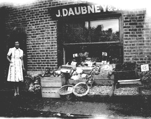 B/W photo taken outside shop, display of fruit and veg in wood boxes and wicker baskets
