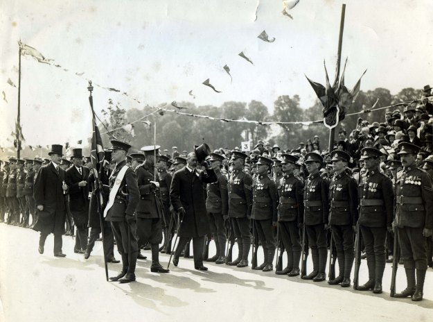 King George V inspecting a line of soldiers of various ranks, with flags and bunting in the background; the public are seated in tiers behind