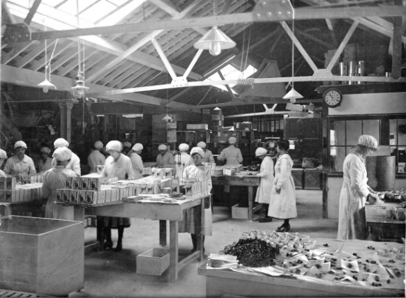 Originally sepia photo of packing room, approaching 20 women in white protective below knee long-armed clothing with white hats (like shower caps) packing goods on wooden tables; the packing room is single storey, no windows but skylights bring in light