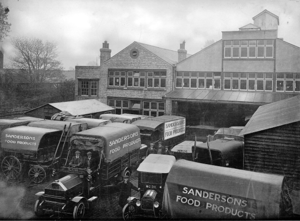 Originally sepia photo from 1921 showing transport yard full of motor lorries and horsedrawn delivery wagons, each with 'Sanderson Food Products' on the side