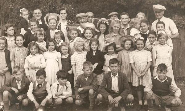 Outdoor sepia photo, around 30 children and 10 adults, the girls in summer dresses