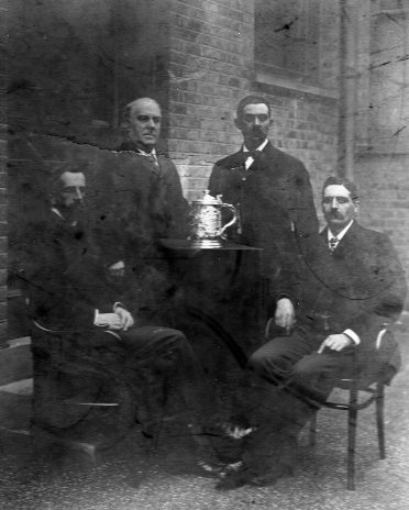 Four men outside, posed around a small table with ornate silver lidded tankard