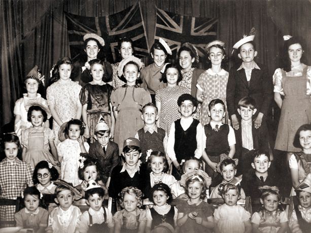 Photo of around 40 children on a stage, from tots to young teens, 2 Union Jacks behind
